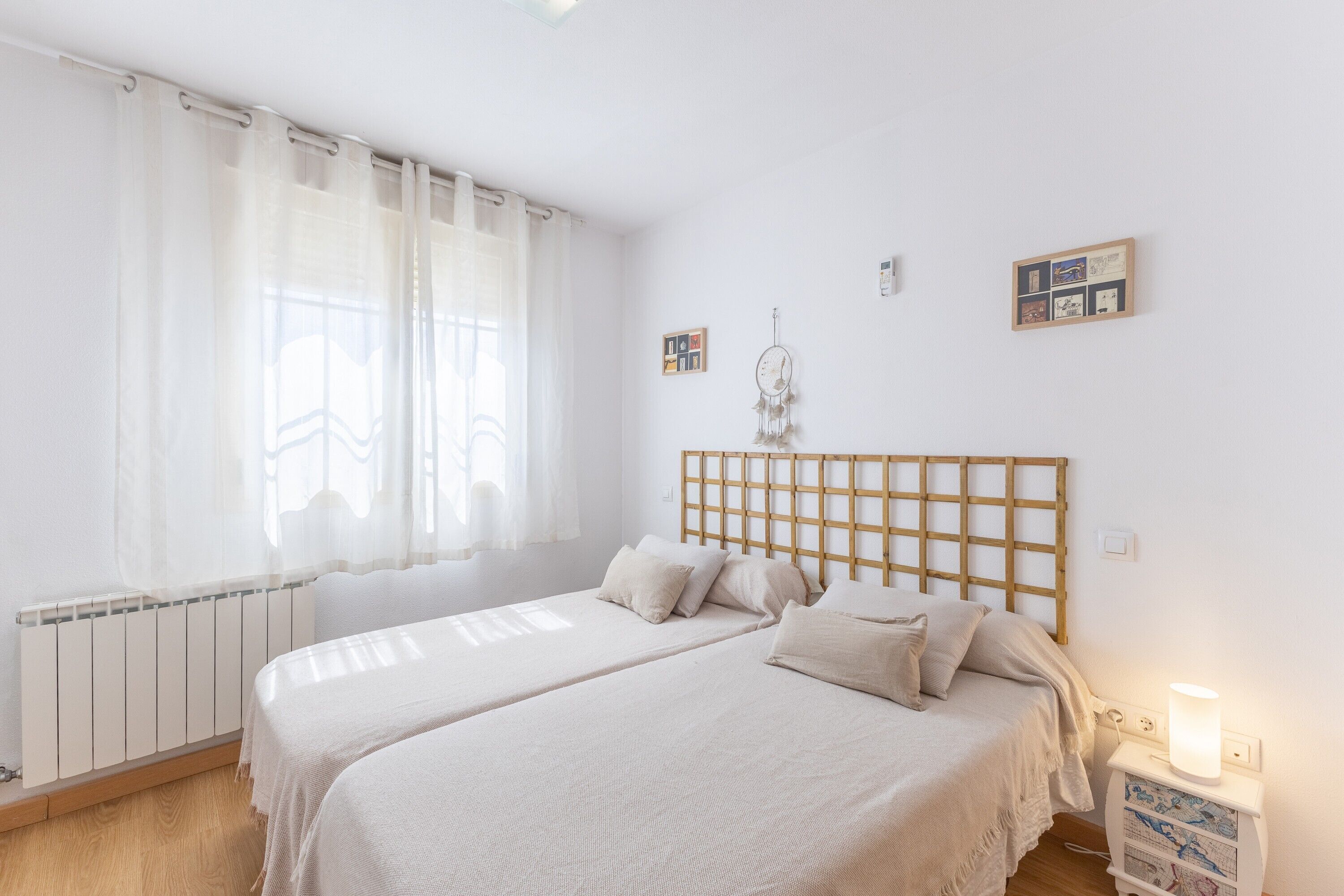 Un dormitorio con dos camas, cabeceros de madera, una mesita de noche con lámpara y una ventana con cortinas translúcidas.