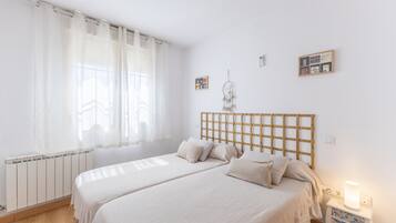 Un dormitorio con dos camas, cabeceros de madera, una mesita de noche con lámpara y una ventana con cortinas translúcidas.