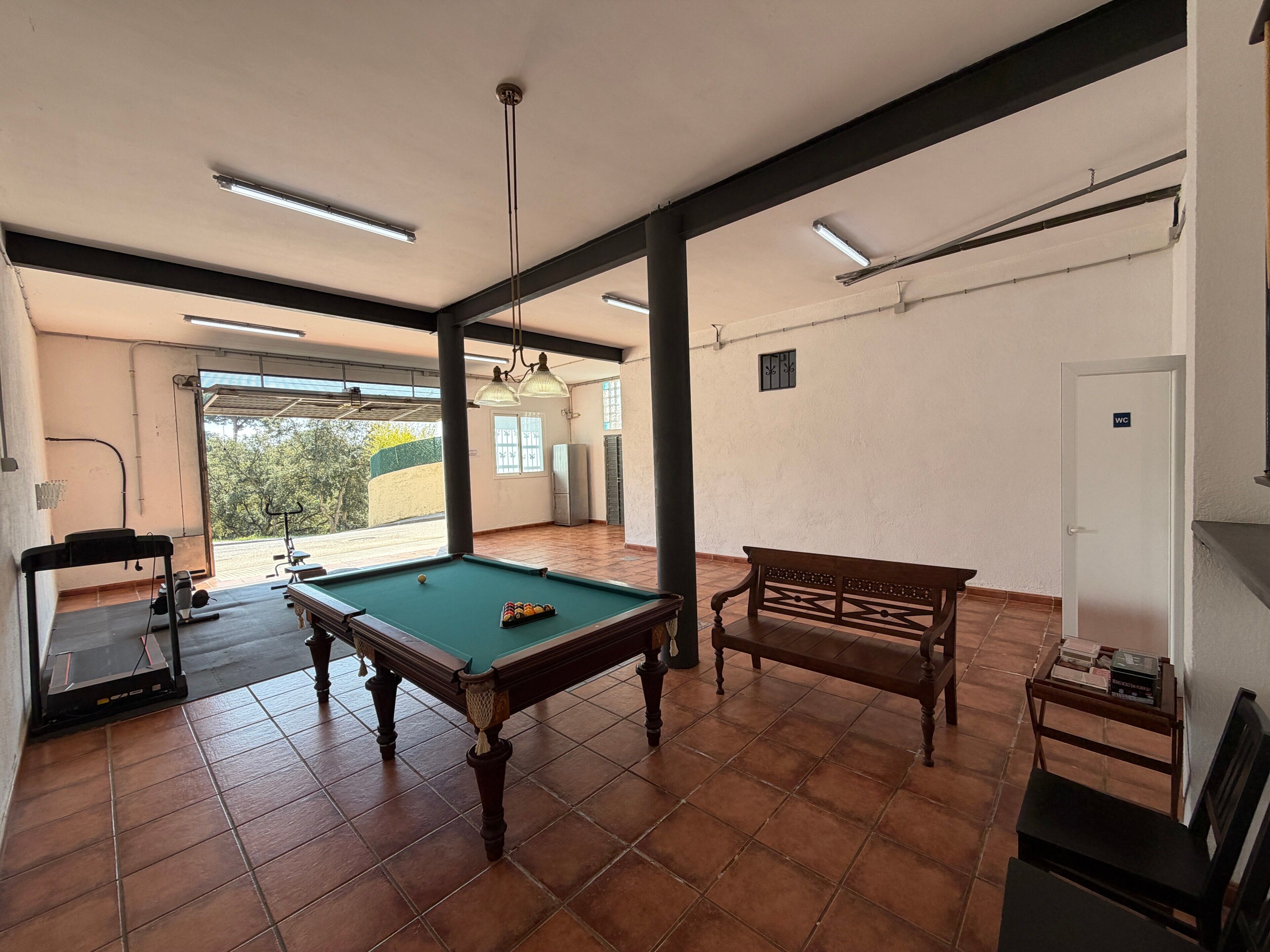 A pool table, a bench, and a door labeled 'WC' in a room with tiled floors and a view through a window.