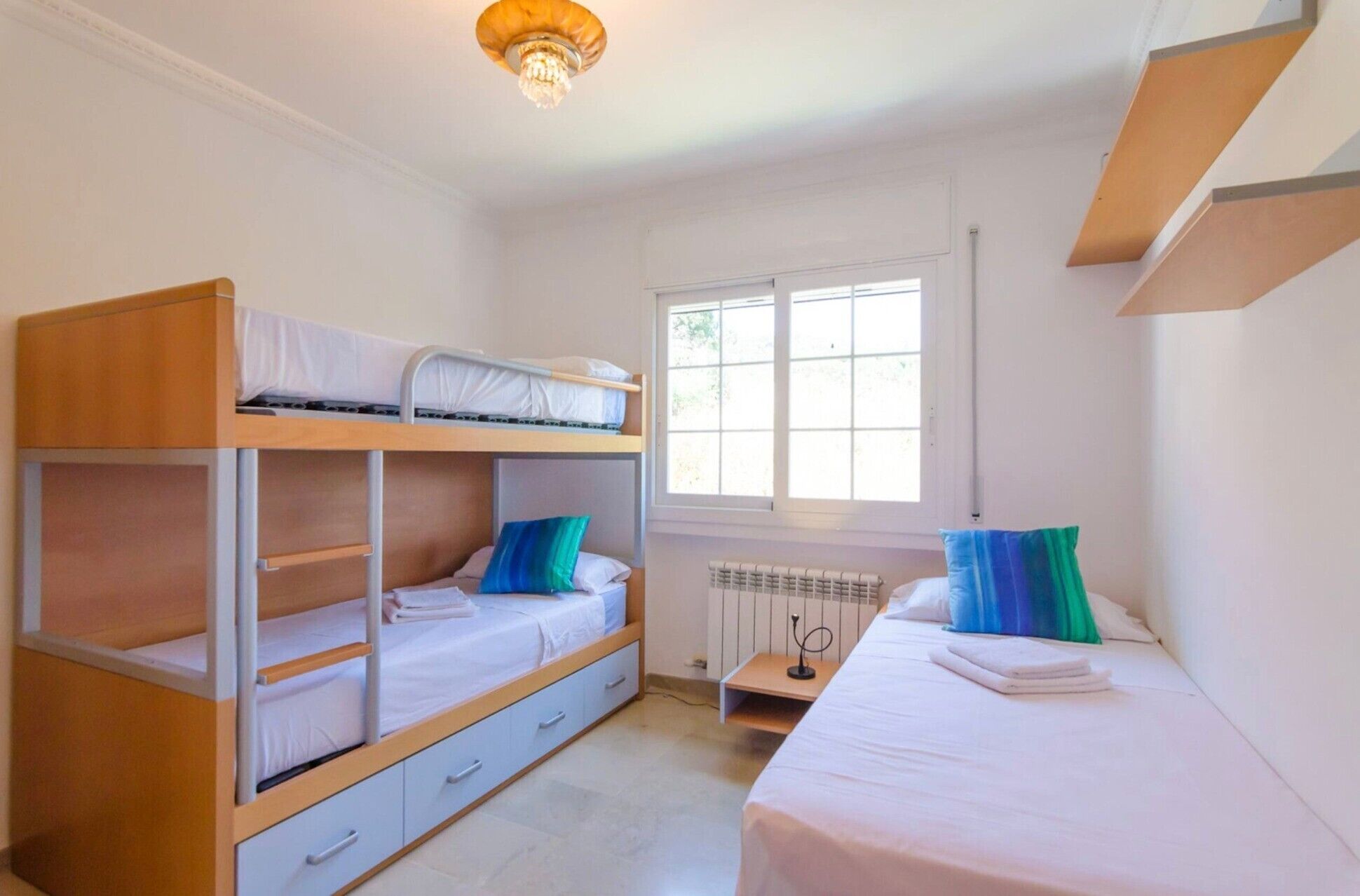 A dormitory room with bunk beds, a window, a radiator, and a small table with a lamp.