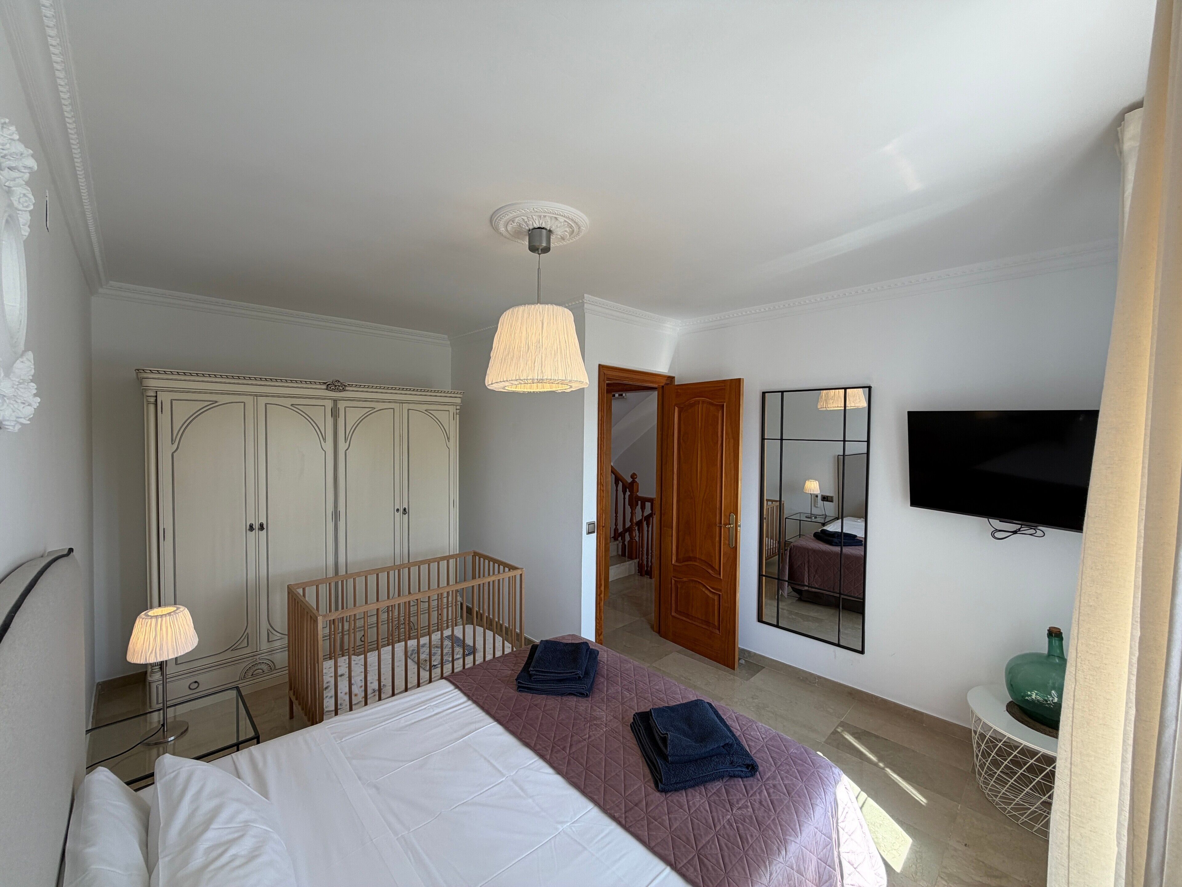 Une chambre à coucher comprenant un lit, un berceau, une armoire, un miroir, une télévision et un escalier menant à une autre pièce.