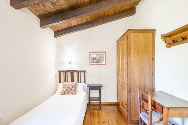 A bedroom with a wooden ceiling, a bed with a patterned pillow, a small table, and a wooden wardrobe.