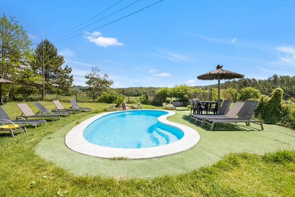 Une piscine entourée de chaises longues et d’une table, le tout bordé de pelouse verdoyante et d’arbres.
