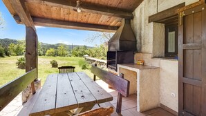 An outdoor patio with a wooden table and benches, a barbecue grill, and a view of the surrounding landscape.