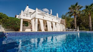 A white building with a balcony, a swimming pool, and palm trees.