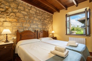 Une pièce avec un mur en pierre, un plafond en bois, deux lits avec du linge de lit blanc, deux lampes de chevet et une fenêtre donnant sur un paysage verdoyant.