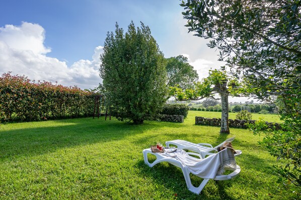 Un espace extérieur verdoyant avec une chaise longue blanche, une table avec une boisson et un grand arbre.