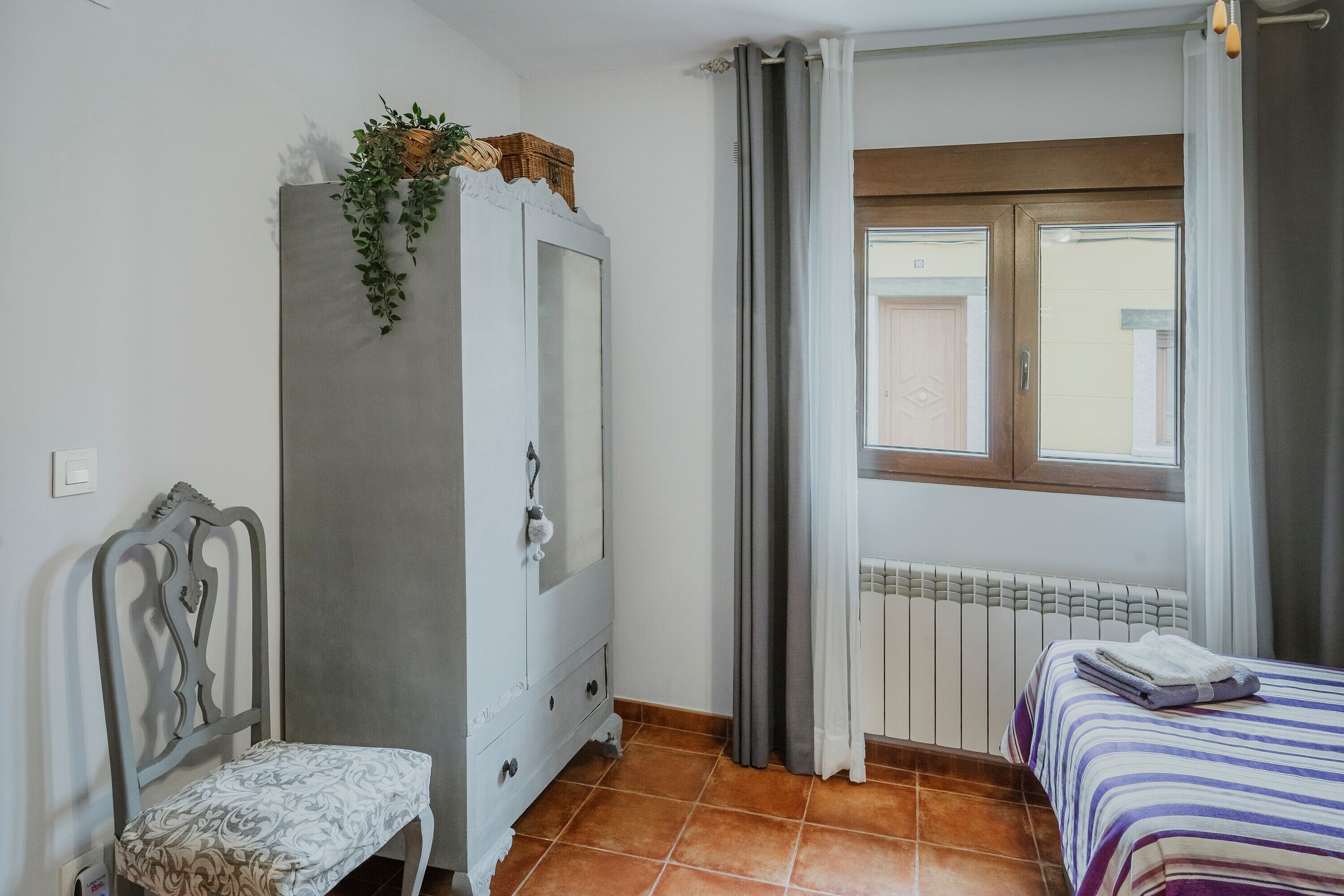Un dormitorio con cama, una silla, armario, ventana con cortinas y radiador.