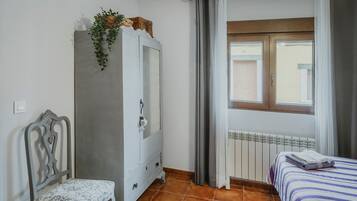 Un dormitorio con cama, una silla, armario, ventana con cortinas y radiador.