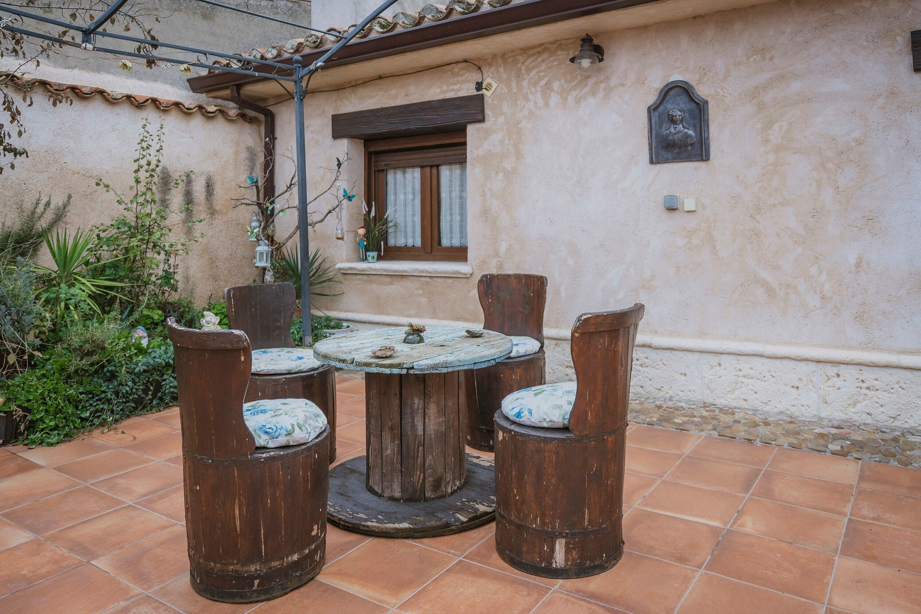 A patio with a round wooden table and four chairs, surrounded by a garden with various plants and a tiled floor.