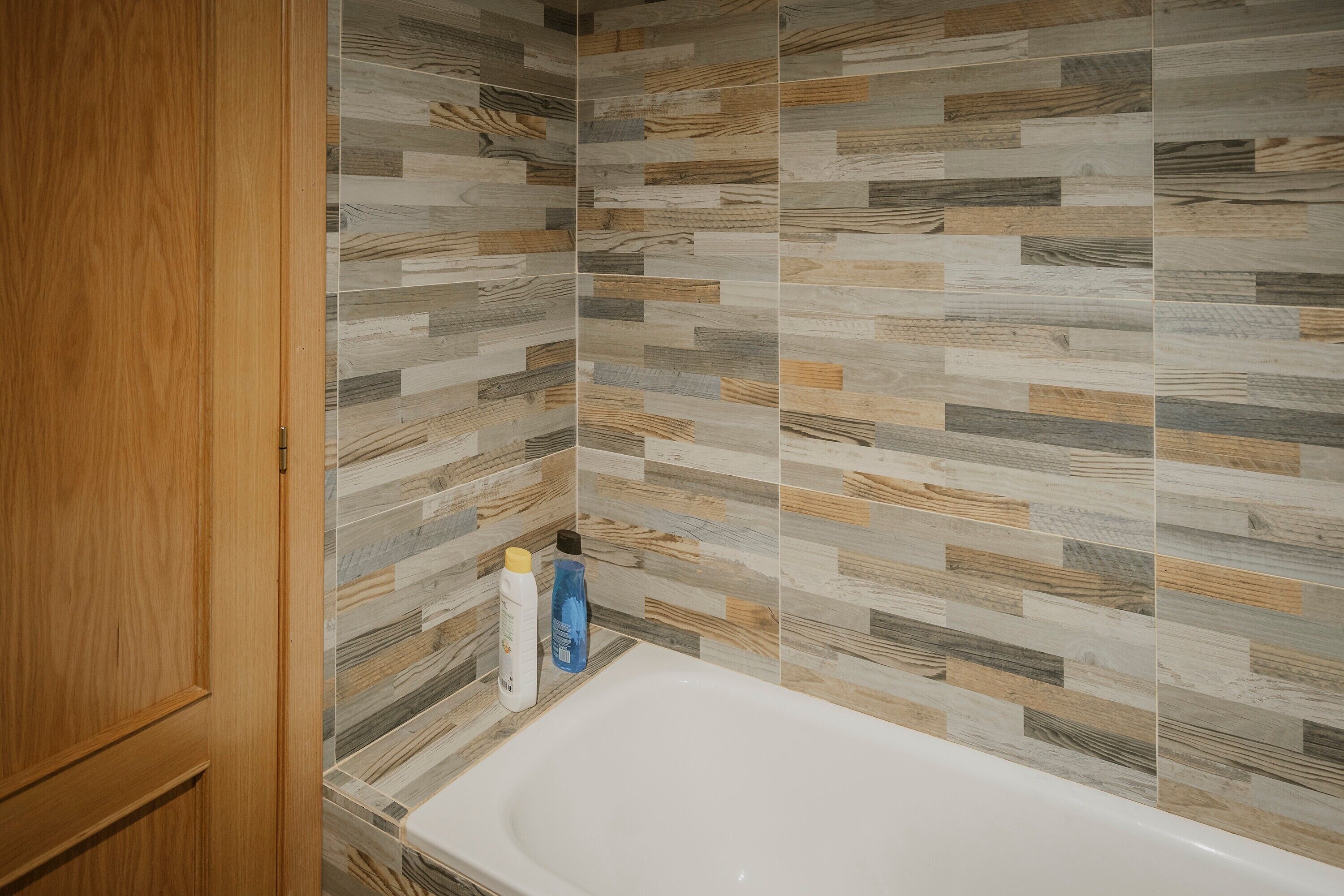 A bathroom with a wooden door, a white bathtub, and a tiled wall with a wood-like pattern.