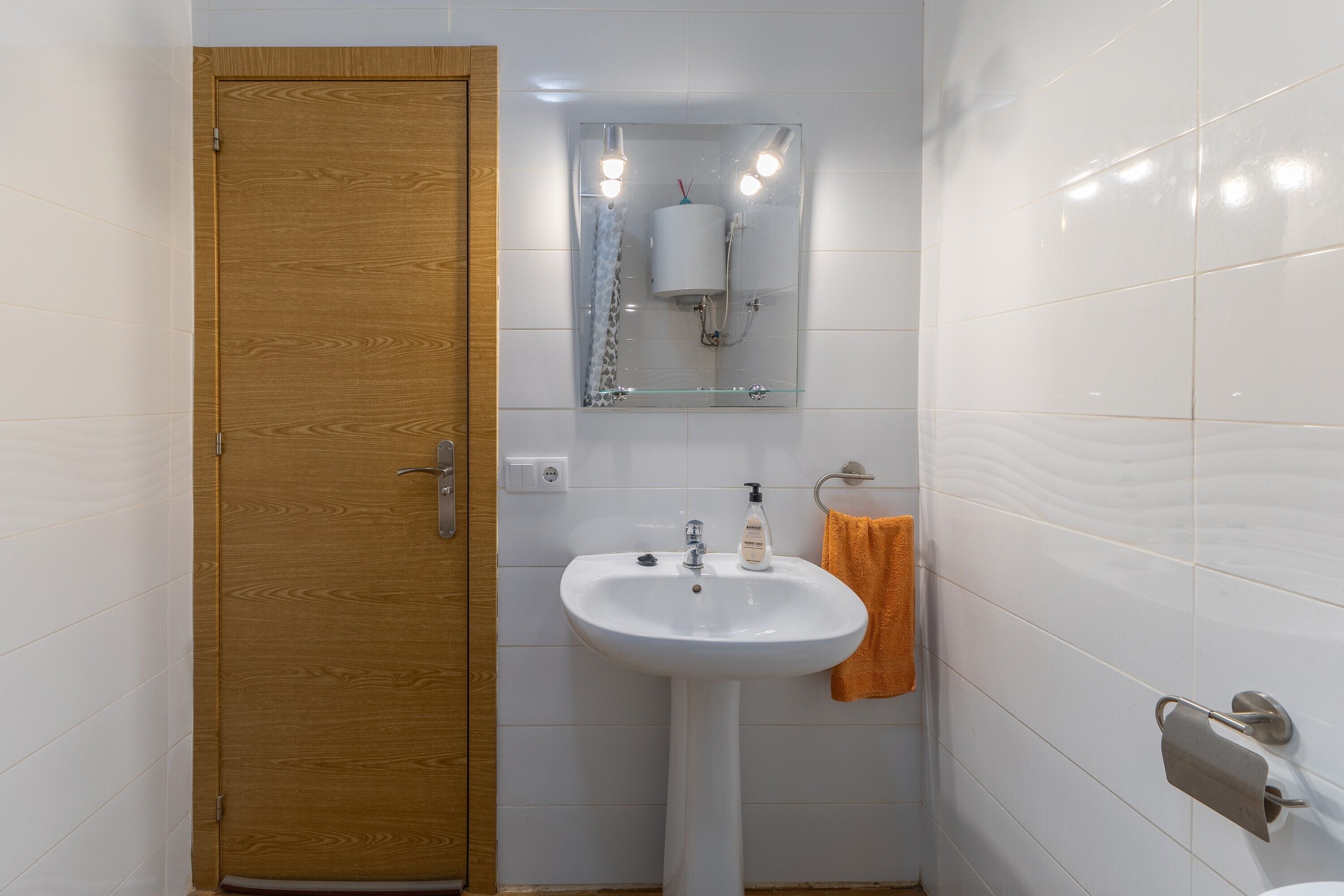 Un bany amb una porta de fusta, un lavabo blanc, un mirall i un suport per a tovalloles.