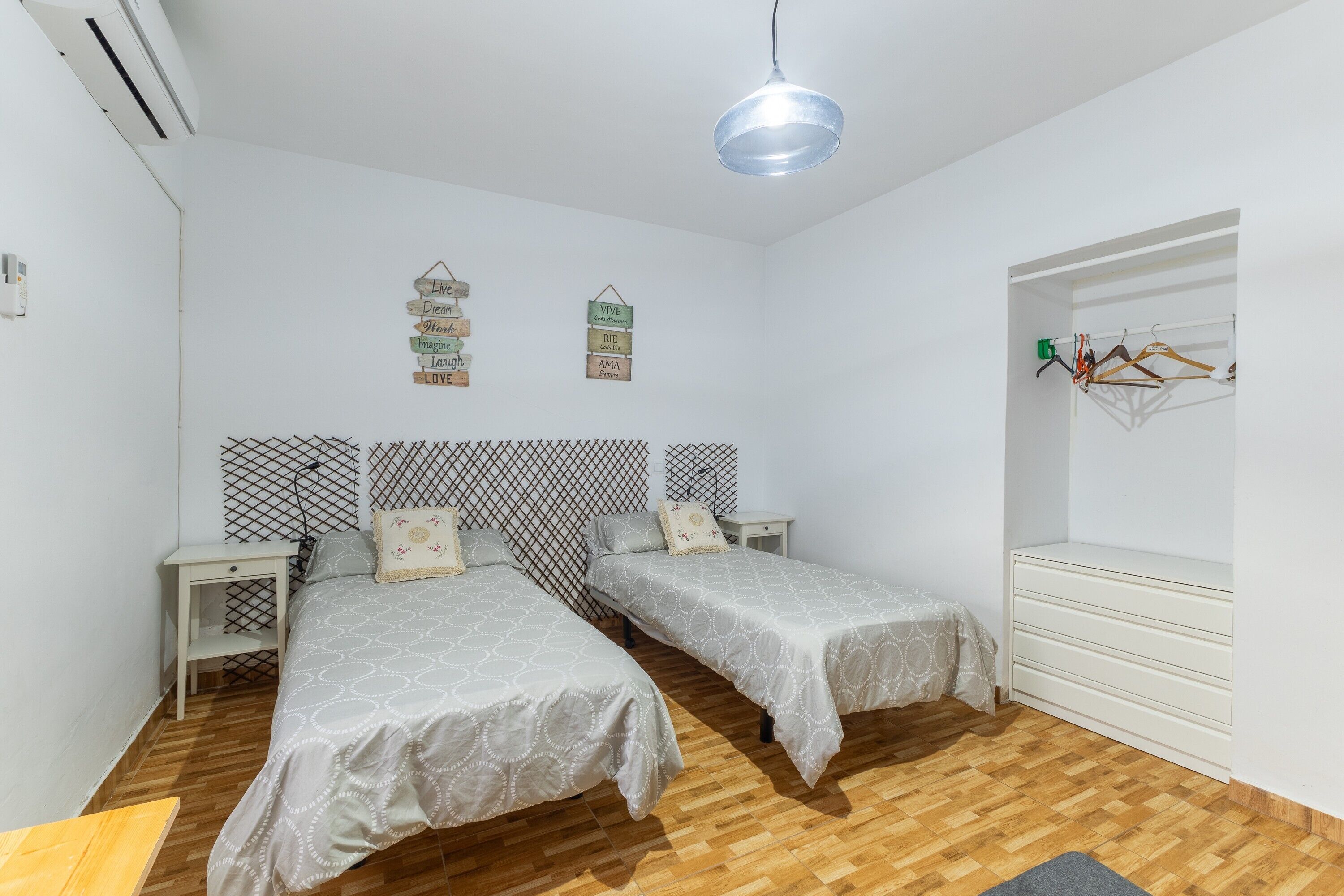 A room with two beds, a wardrobe, a bedside table, a hanging light fixture, and wall decorations.