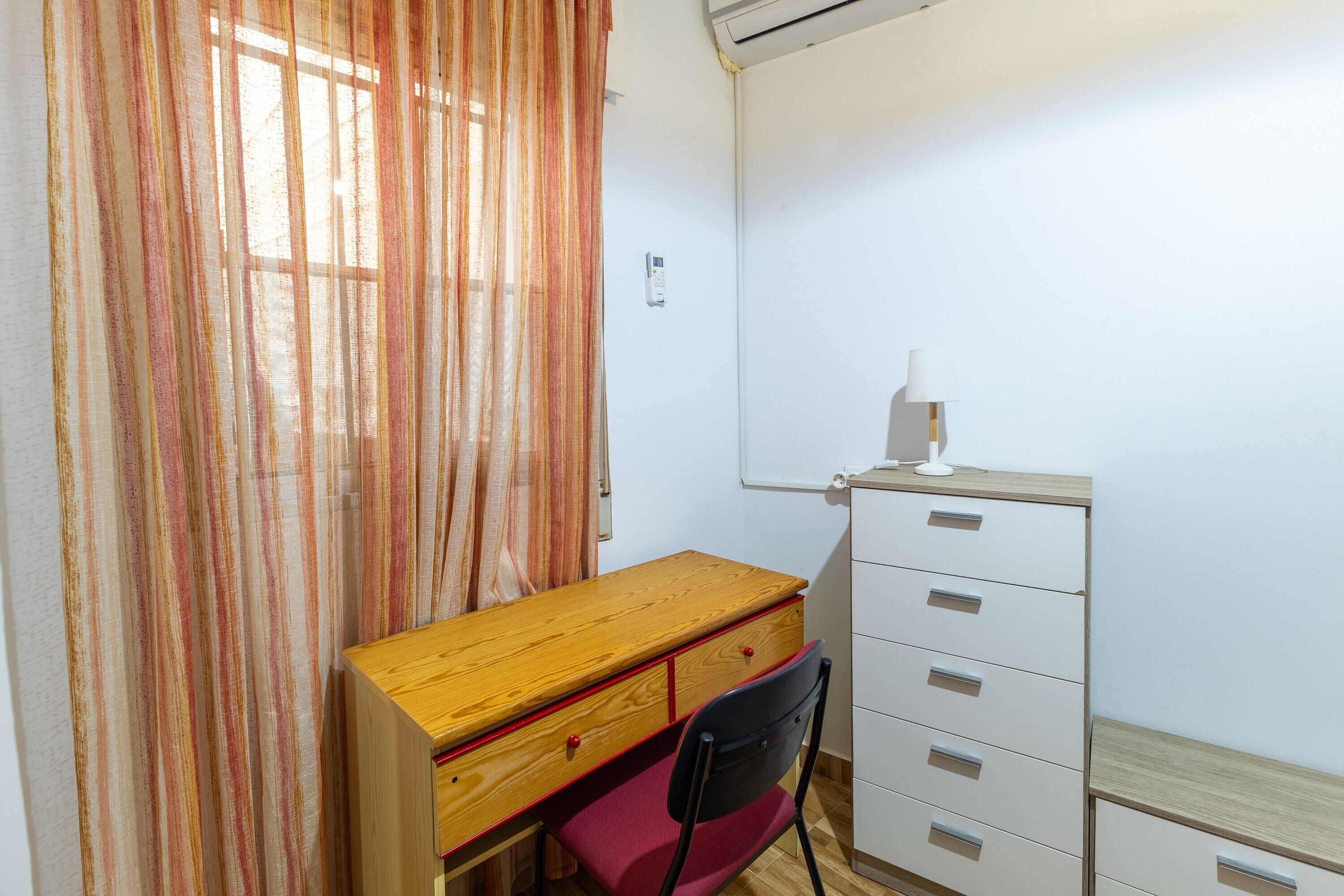 Una habitación con un escritorio de madera, una silla roja, un archivador blanco y un ventanal con cortinas.