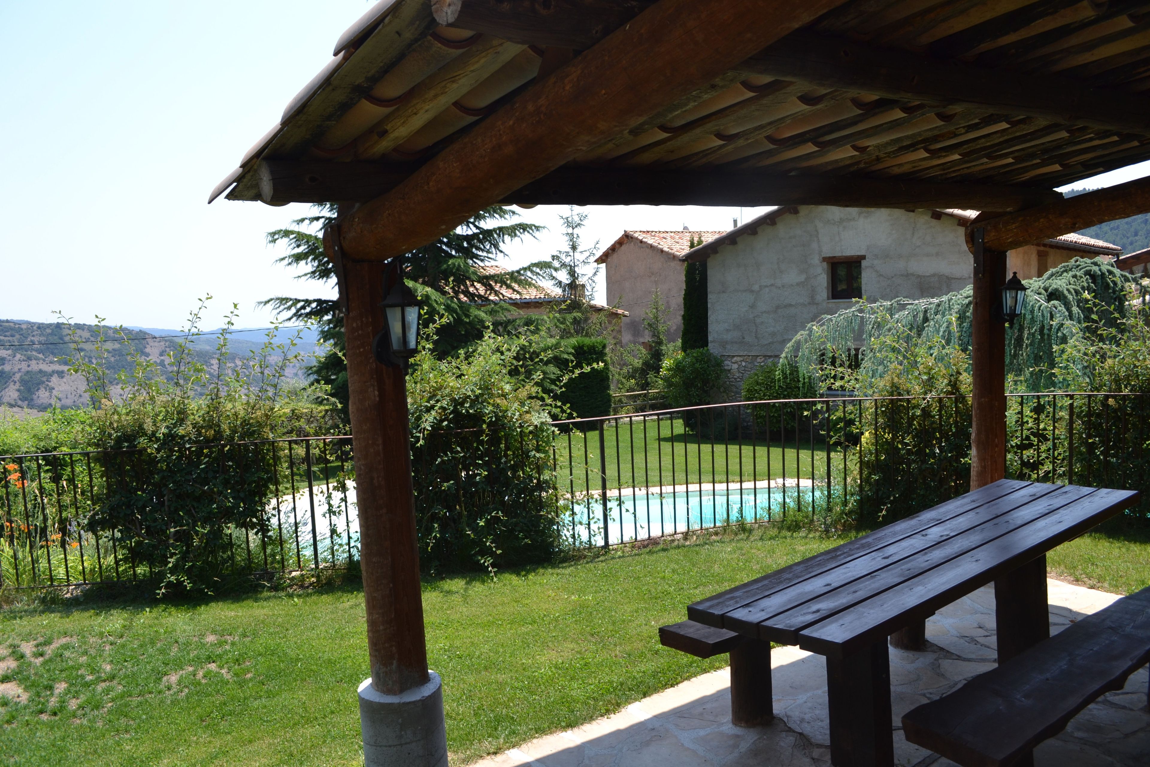Una pérgola de madera con un banco, con vistas a una piscina y un área verde con vistas a colinas distantes.