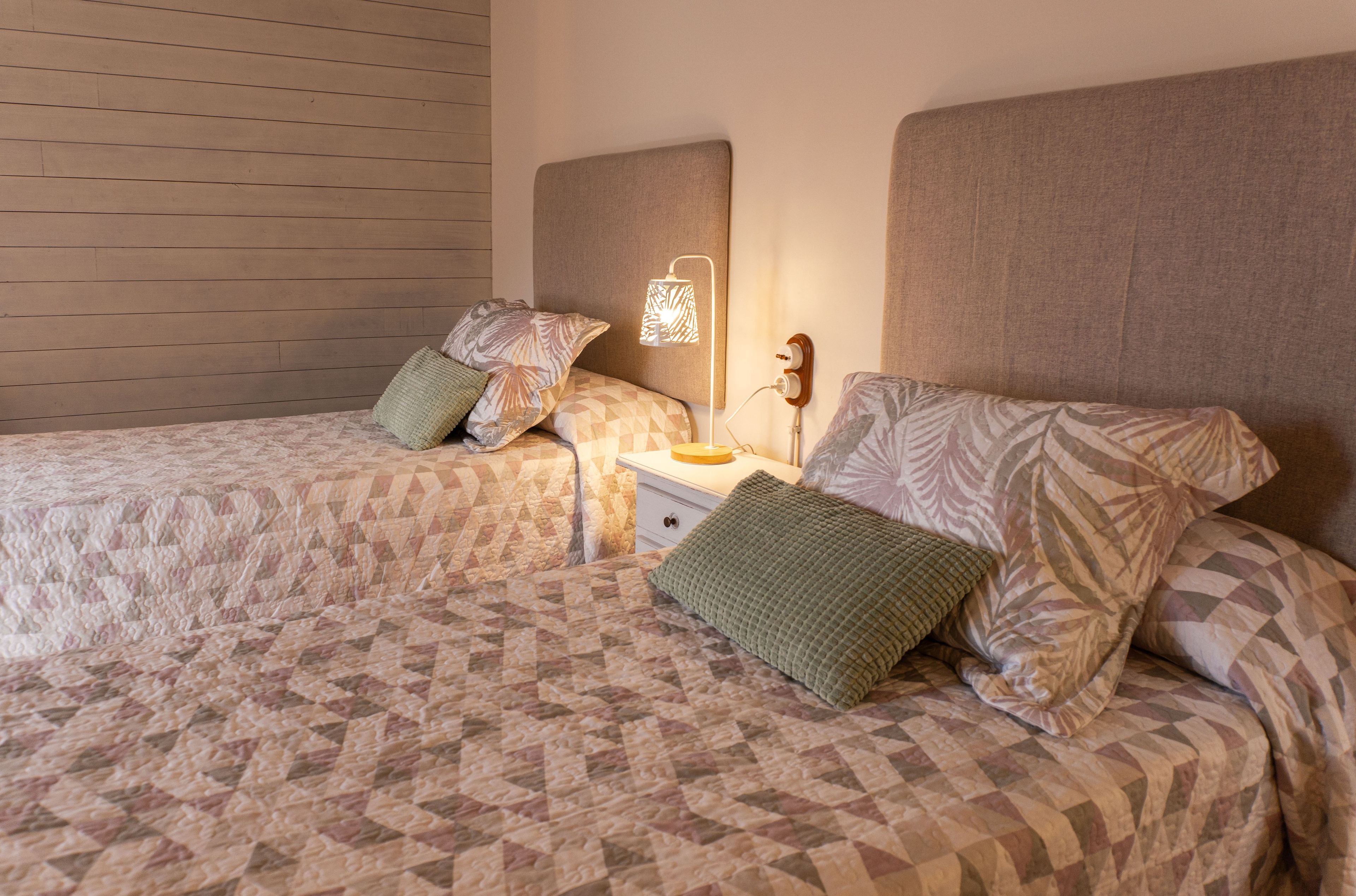 Een tweepersoonsbed met een geborduurde sprei, twee kussens, een nachtkastje met een lamp, en een wand met horizontale houten panelen.