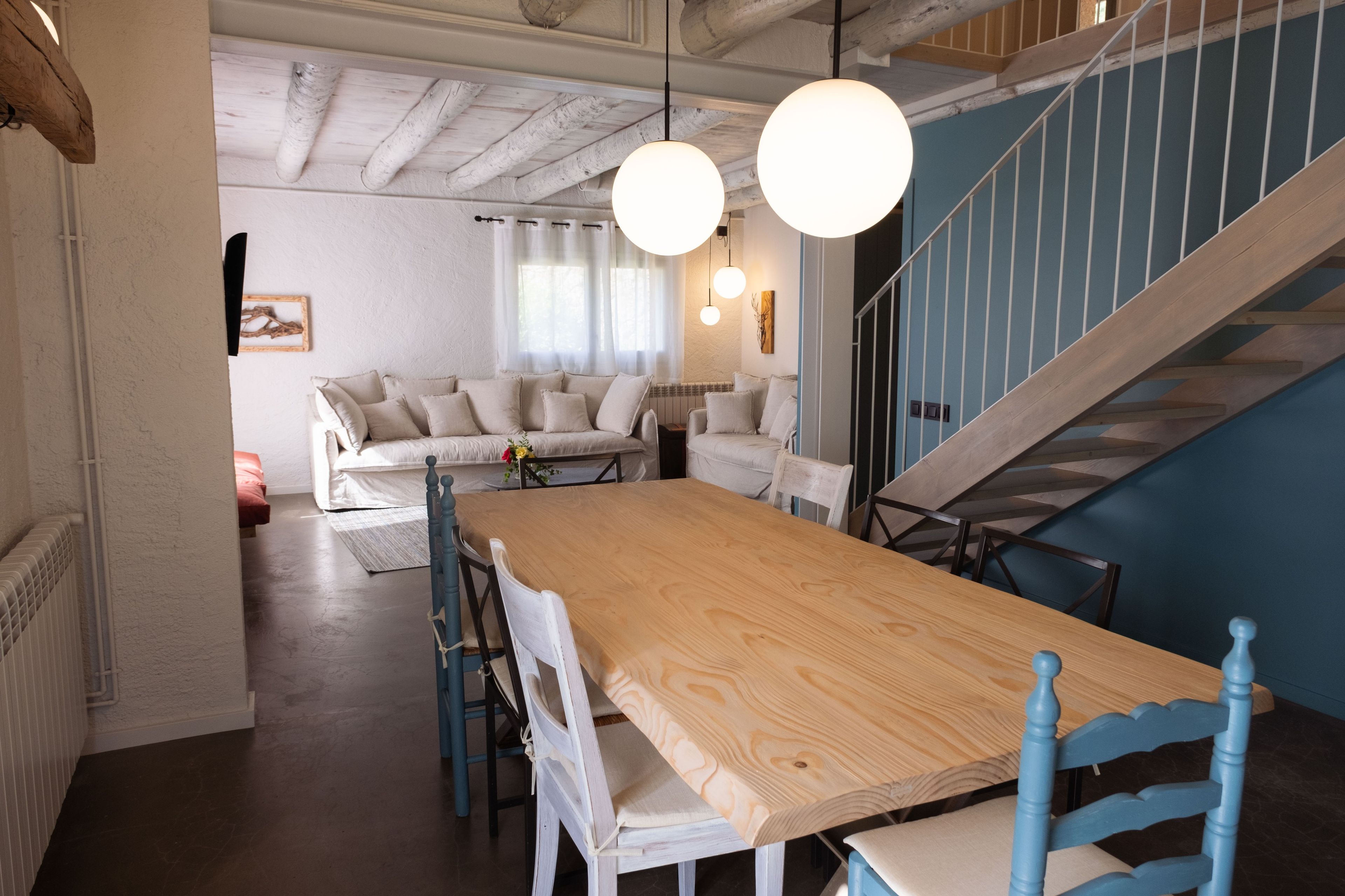 A dining area with a long wooden table and chairs, a staircase, and a living room with a sofa and curtains.