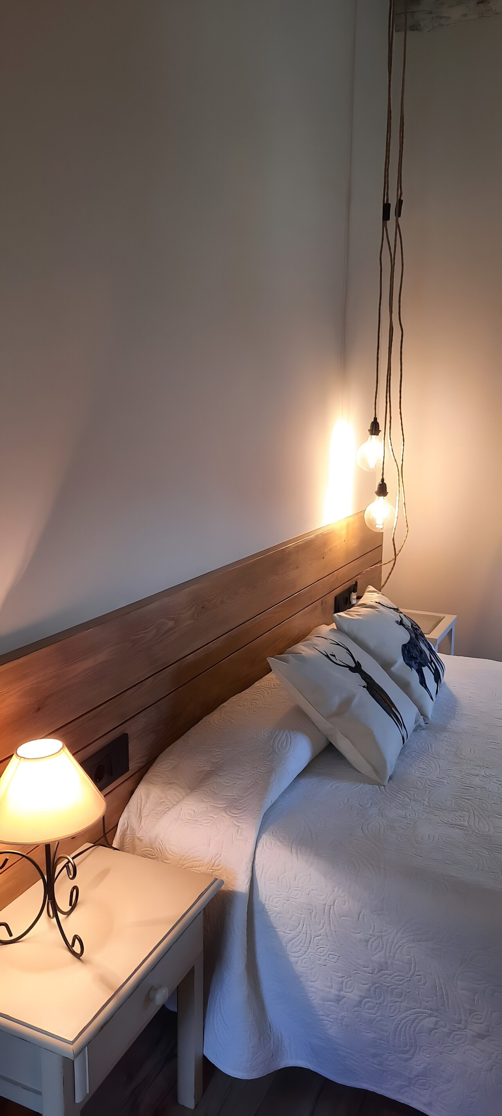 A neatly made bed with a wooden headboard, a bedside table with a lamp, and hanging light bulbs.