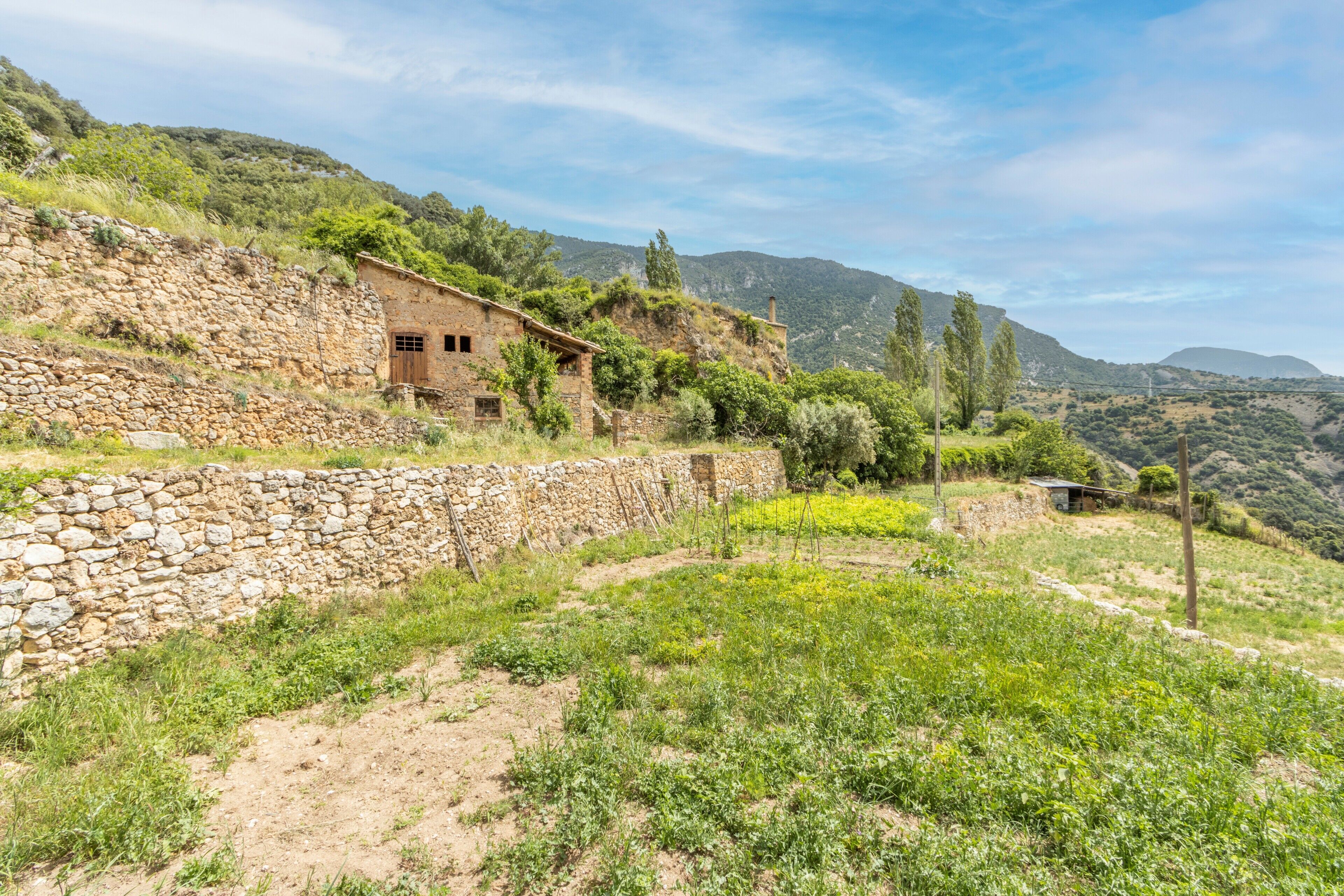 A stone house with a dirt path, surrounded by greenery and hills.