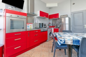 Een kleine keuken met rode keukenkastjes, een ingebouwde oven, magnetron en wasmachine. Er staat een eettafel met een blauw-witte tafelkleed.