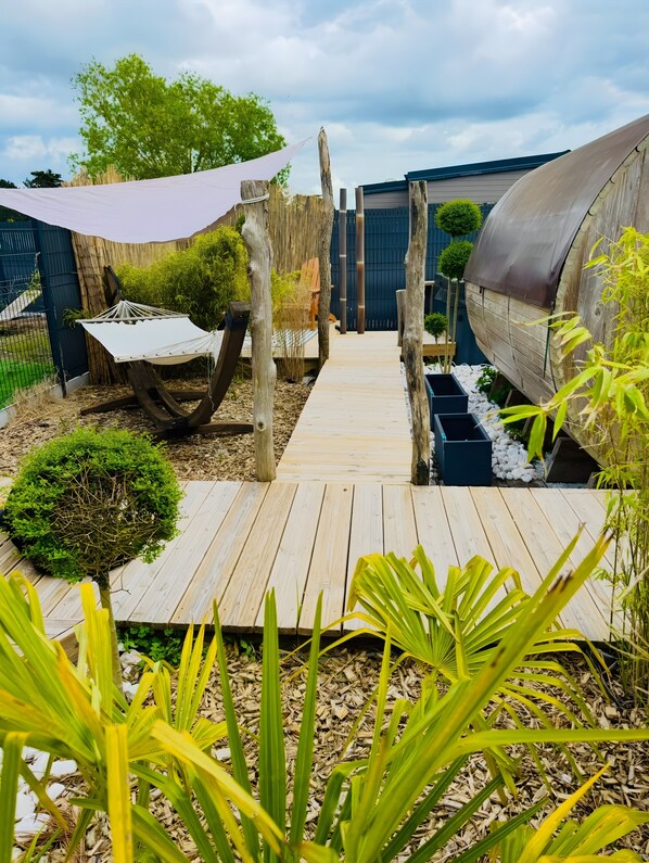 A wooden deck leading to a covered outdoor area with a hammock, a small garden, and a wooden structure.