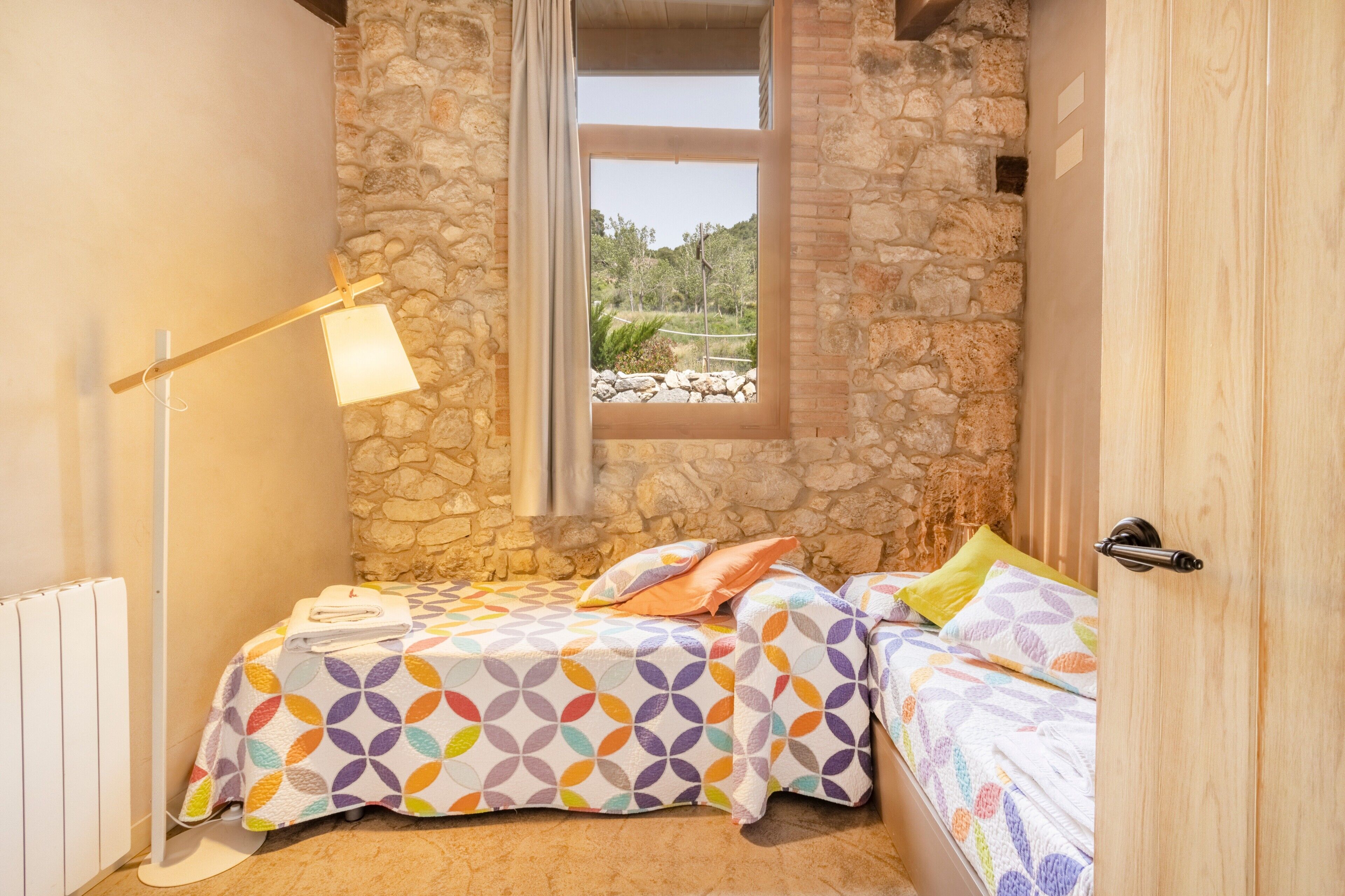 A bedroom with a stone wall, a window with a view, a bed with patterned bedding, and a floor lamp.