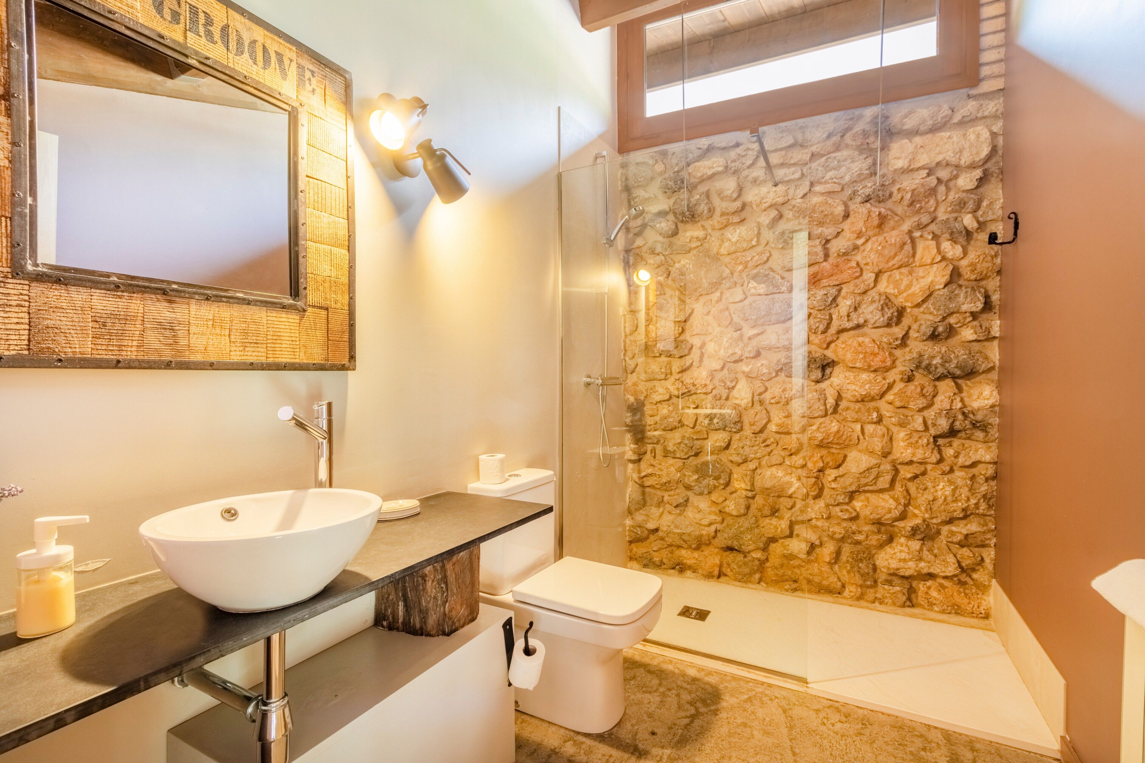 Een badkamer met een stenen muur, een witte wastafel, een toilet en een douche met glazen wanden.