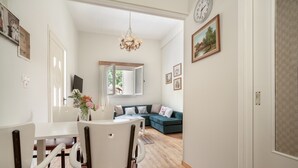 A dining area with a white table and chairs, a chandelier, and a view into a living room with a sofa and a window.