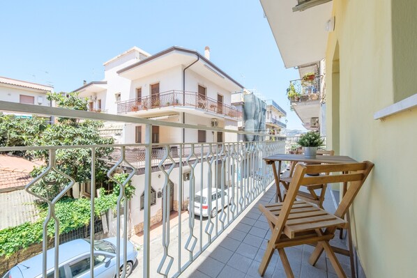 A balcony with a wooden table and chairs, a metal railing, and a view of neighboring buildings and parked cars.