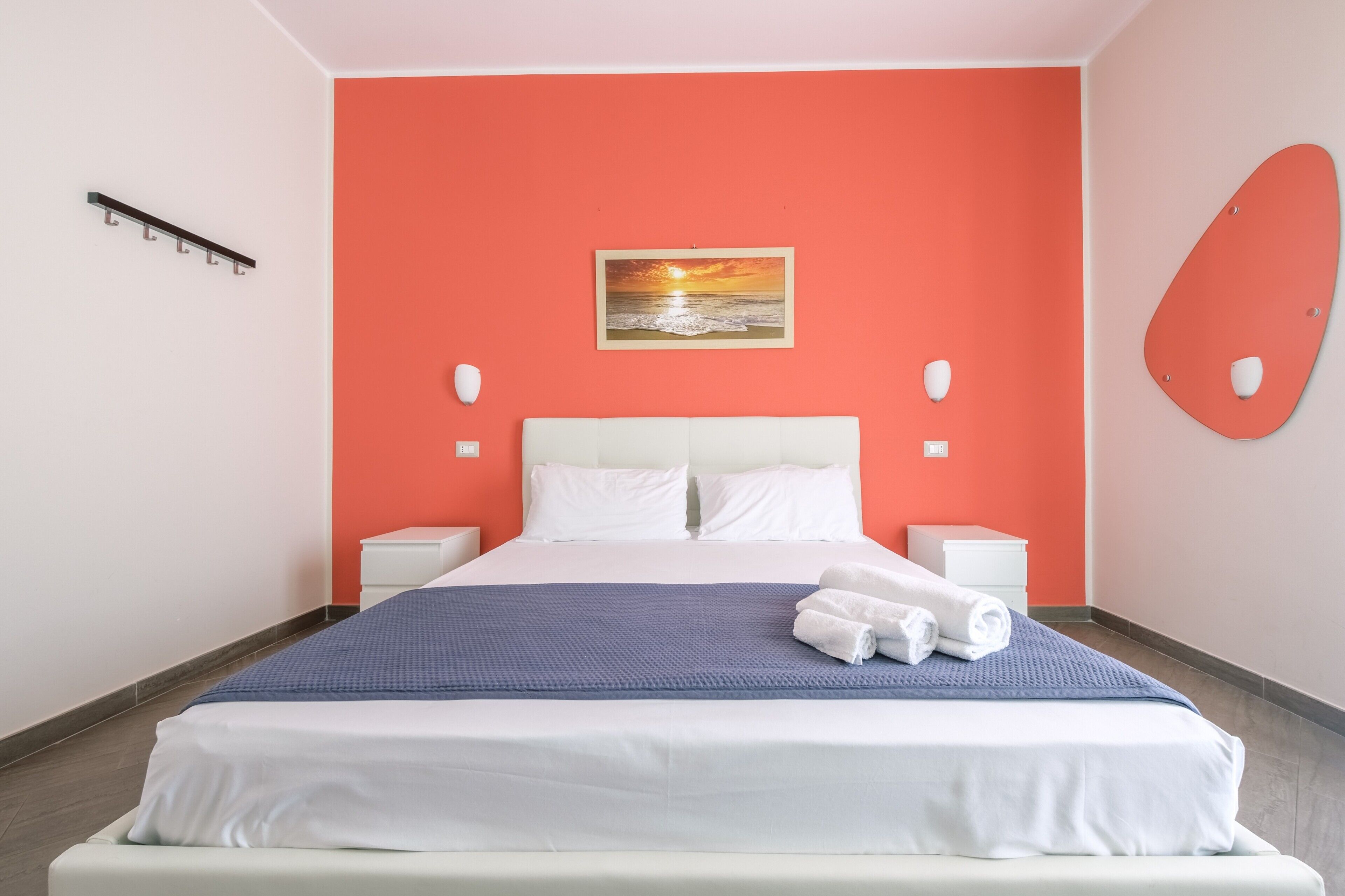 A hotel room with a bed, white nightstands, a blue blanket, and a sunset painting on the wall.