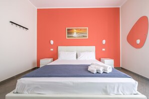 A hotel room with a bed, white nightstands, a blue blanket, and a sunset painting on the wall.