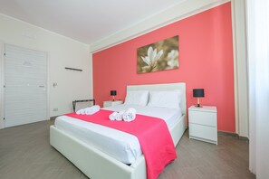Une chambre d’hôtel avec un lit, des murs blancs, un mur d’accent rouge, un tableau, une table de chevet, une lampe et une porte fermée.