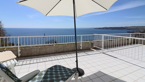 Une terrasse dotée d’un parasol blanc, d’une balustrade blanche et offrant une vue sur la mer.