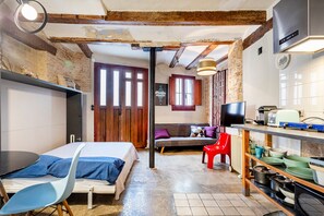 A room with a bed, a red chair, a sofa, a TV, and a kitchen area with a stove and shelves.