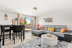 Un salon avec un canapé gris, une table basse ronde, une table à manger avec des chaises, et une vue sur l’extérieur à travers une porte vitrée.