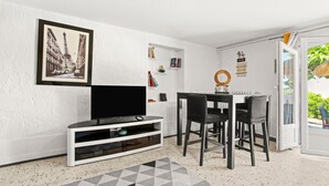 Una habitación con un televisor de pantalla plana sobre un soporte blanco, una mesa de comedor con sillas, una estantería y una puerta que da a un espacio exterior.