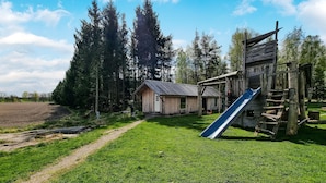 Una baita rustica in legno con tetto in metallo, una piccola rimessa in legno e uno scivolo blu in un'area erbosa con alberi sullo sfondo.