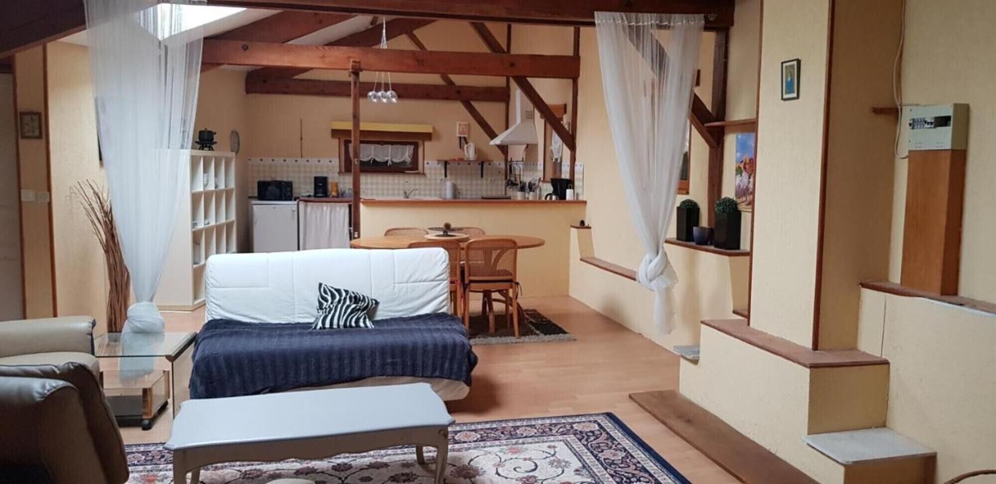 Una habitación acogedora con cama, una mesita, una silla, un sofá, y una zona de cocina al fondo, además de una escalera.