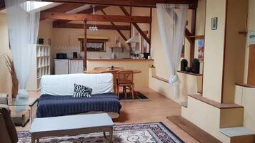 Una habitación acogedora con cama, una mesita, una silla, un sofá, y una zona de cocina al fondo, además de una escalera.