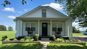 A white single-story house with a front porch, two rocking chairs, and a small garden area. - Cozy modern farmhouse w/ garage- Only 5 miles to Lake Cumberland! (Monticello)