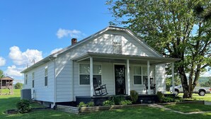 A single-story white house with a front porch, a chimney, and a small garden area. - Cozy modern farmhouse w/ garage- Only 5 miles to Lake Cumberland! (Monticello)