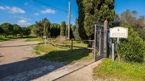 Una entrada rural con una verja de madera, un cartel que dice 'Agriturismo Rosaspina' y un camino de grava que se adentra en la vegetación.