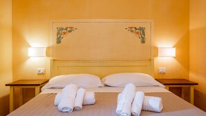 Una cama bien hecha con sábanas blancas y toallas enrolladas, dos lámparas de noche y un adorno floral en la pared.