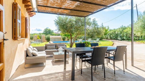 Un patio exterior con una mesa y sillas, un sofá, y vistas a una piscina y vegetación.