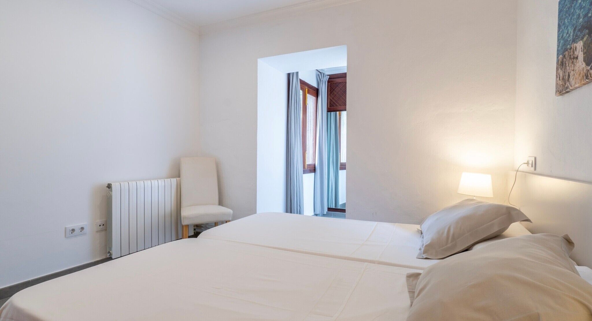 Une chambre d’hôtel avec un lit, une chaise, un radiateur, une fenêtre avec des rideaux et un tableau au mur.