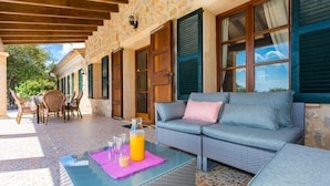 A covered outdoor seating area with a glass table, wicker furniture, and a stone wall. - Country House 'Es Corto' with Mountain View, Private Pool and Wi-Fi (Illes Balears)