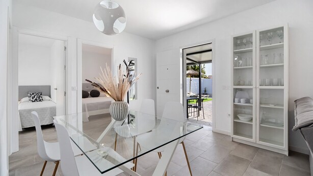 A modern dining area with a glass table and white chairs, a white cabinet with glass doors, and a view to the outside.