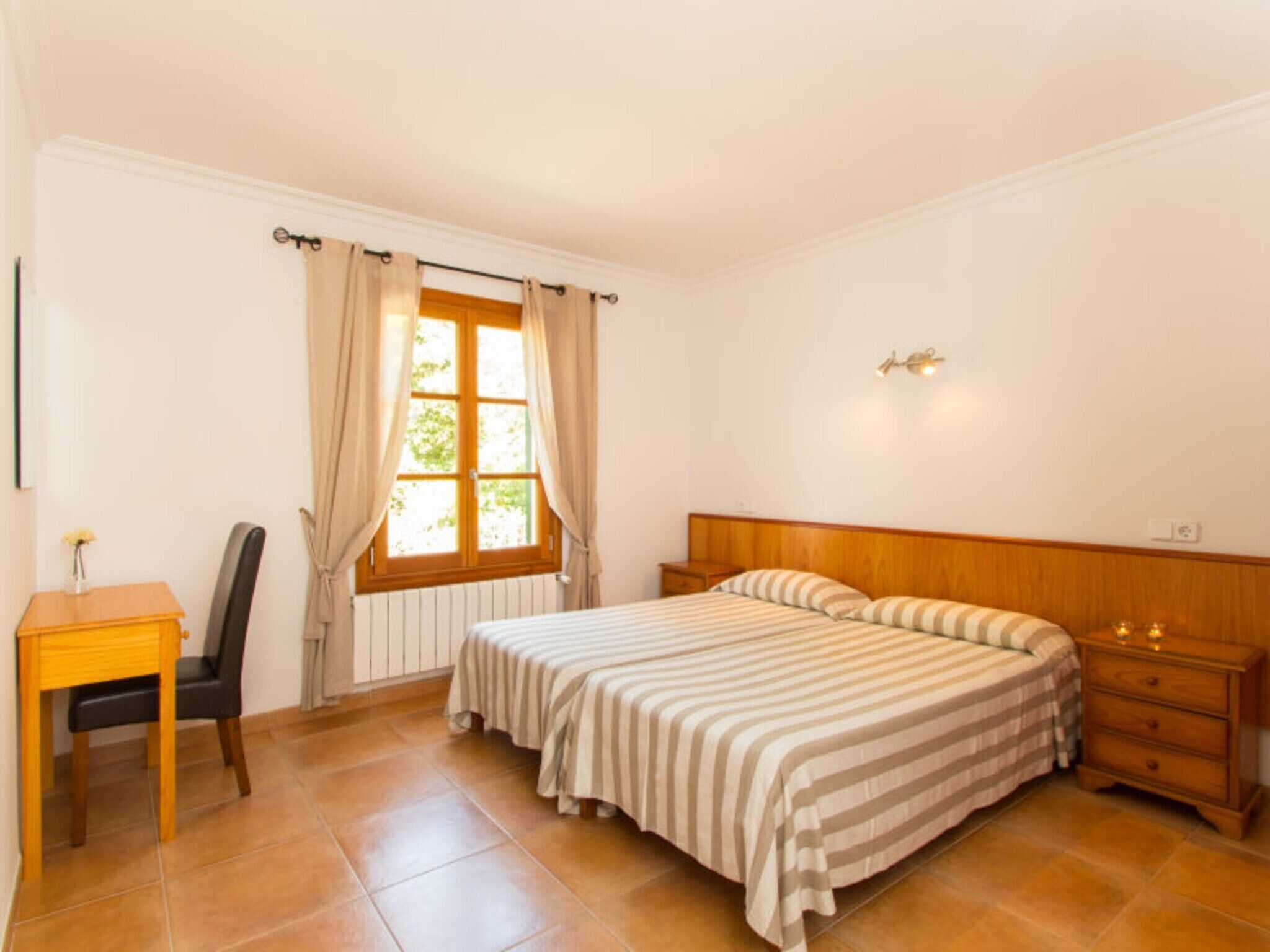 A bedroom with two beds, a desk, a chair, a window with curtains, and a nightstand with a lamp.