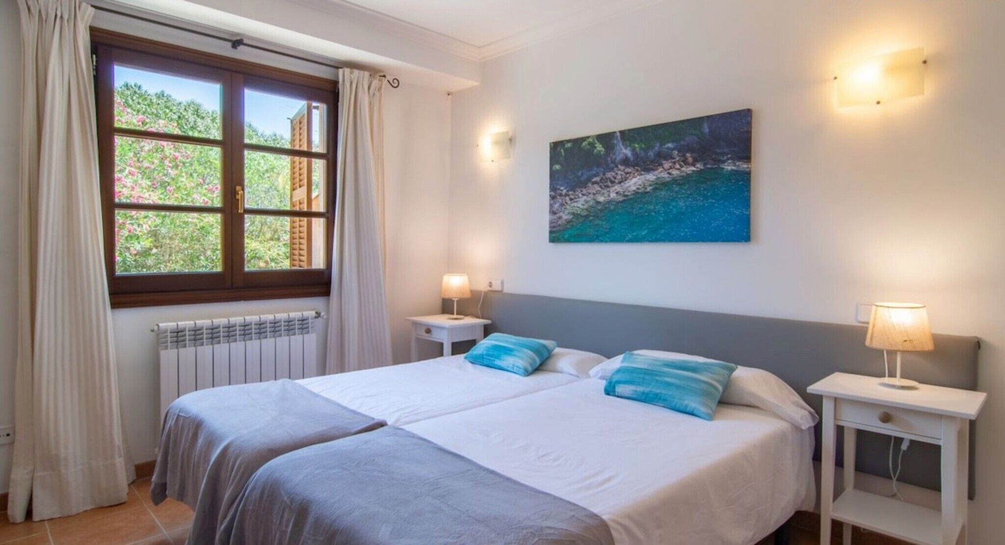 Un dormitorio con una cama, dos mesitas de noche, una ventana con cortinas, un radiador y un cuadro en la pared.