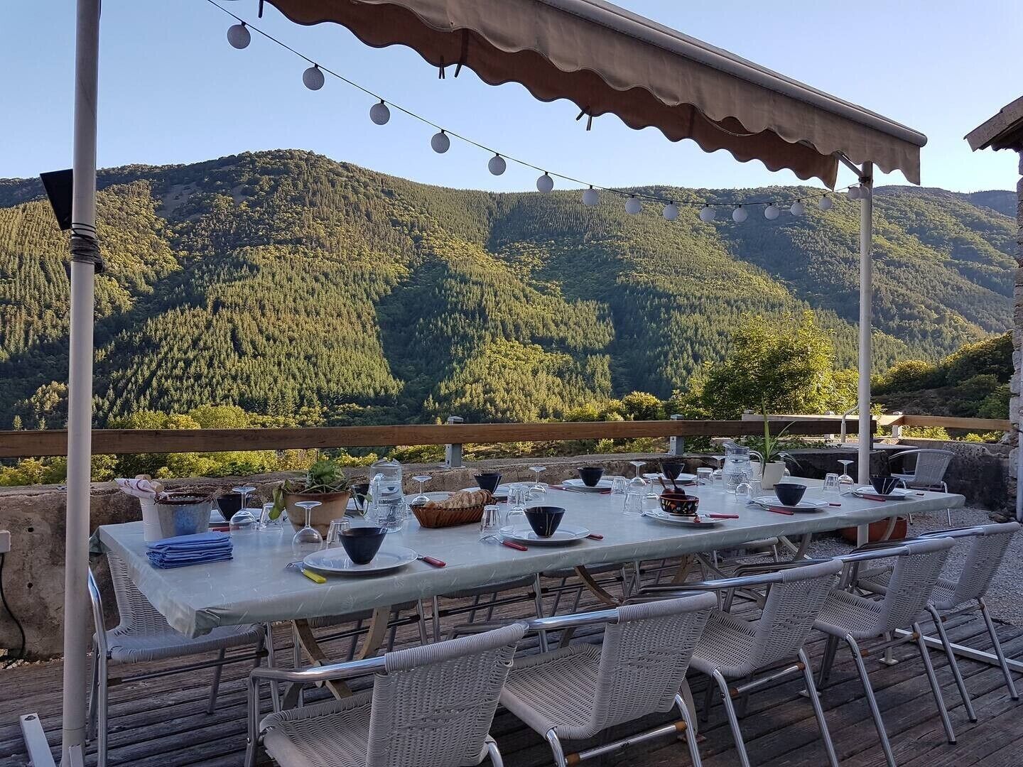 An outdoor dining area with a table set for four, surrounded by wicker chairs, overlooking a forested mountain range.